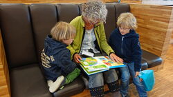 Schulbibliotheken in Kooperationsschulen der tandem BTL Lesepaten: Frau liest zwei Kindern vor, in der Schulbibliothek